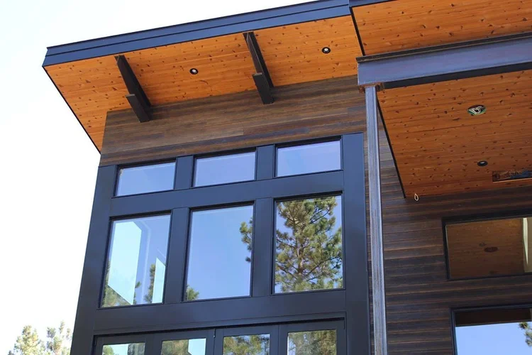 Close-up of a modern house exterior with large glass windows, wooden panels, and a wooden ceiling with black supporting beams.