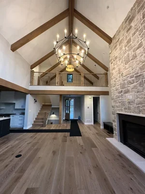 Empty living room with high vaulted ceiling, chandelier, stone fireplace, staircase, and upper balcony overlooking the space.