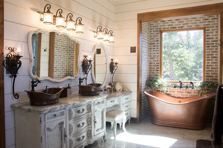 A rustic bathroom with a double vanity with two vessel sinks and ornate mirrors, a vintage style wood cabinet, and a copper clawfoot bathtub near a large window with a brick wall background and green plants.