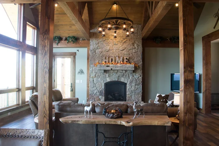 Living room with a stone fireplace, wooden beams, and a chandelier, decorated with a wooden word sign 'HOME', and furniture including armchairs, a television, and a rustic wooden table.