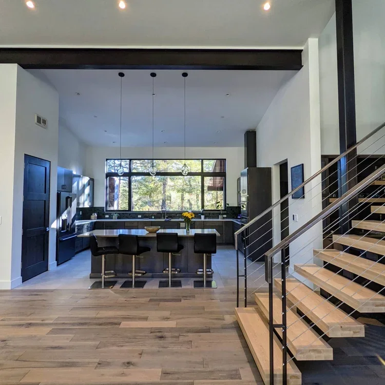 Modern open-concept kitchen with large window, black cabinets, center island with four black chairs, and a staircase with wooden steps and metal railing.