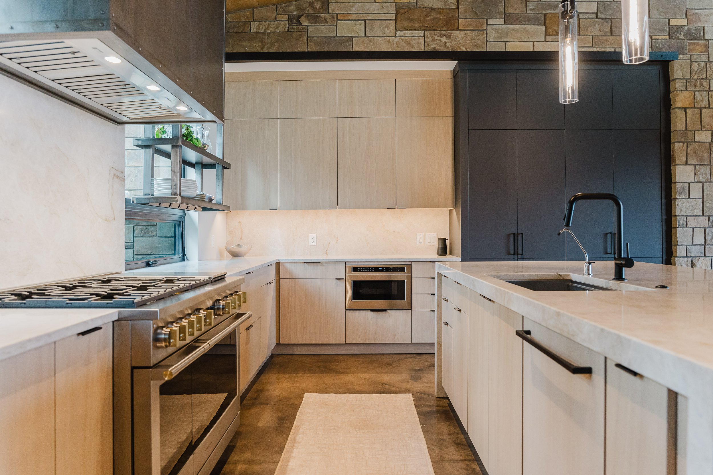Modern kitchen with beige cabinets, stone wall, black upper cabinets, marble countertops, stainless steel appliances including a stove and oven, black sink with a modern faucet, pendant lights, and a window with plants outside.