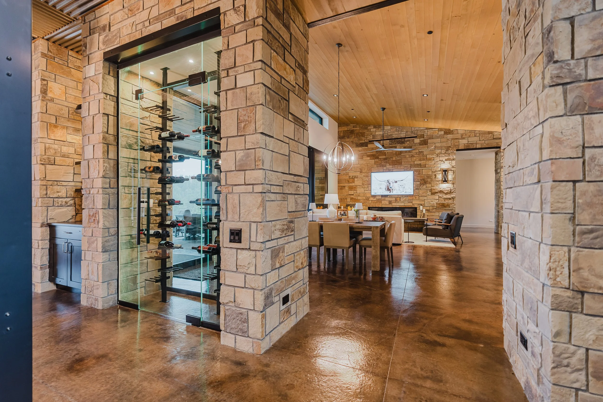 Interior of a room with stone walls, wooden ceiling, and polished concrete floor. There is a glass wine storage display with bottles and a dining area with chairs, a table, and a TV on the wall.