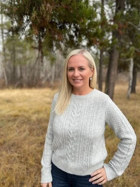 A woman with long blonde hair standing outdoors in front of trees, wearing a light gray sweater and dark jeans, smiling at the camera.