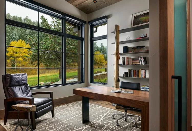 Home office with large window, wooden desk, black office chair, leather armchair, bookshelf, and view of trees outside.