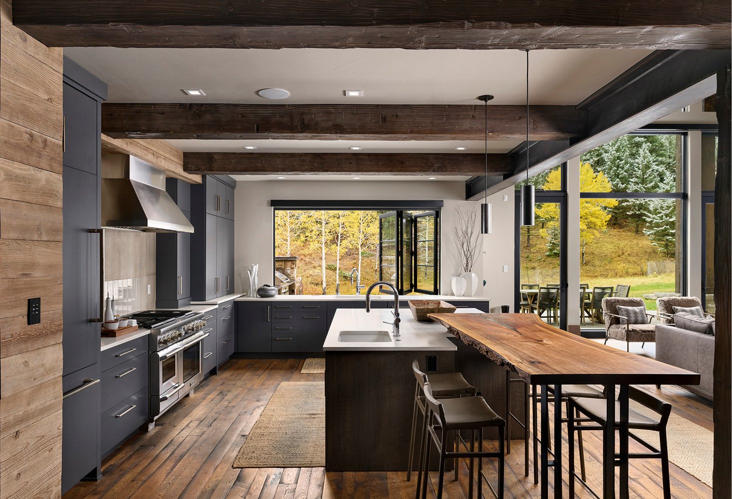 Modern kitchen with dark gray cabinets, white countertops, and a rustic wooden island, featuring large windows with a view of a wooded yard with fall foliage.