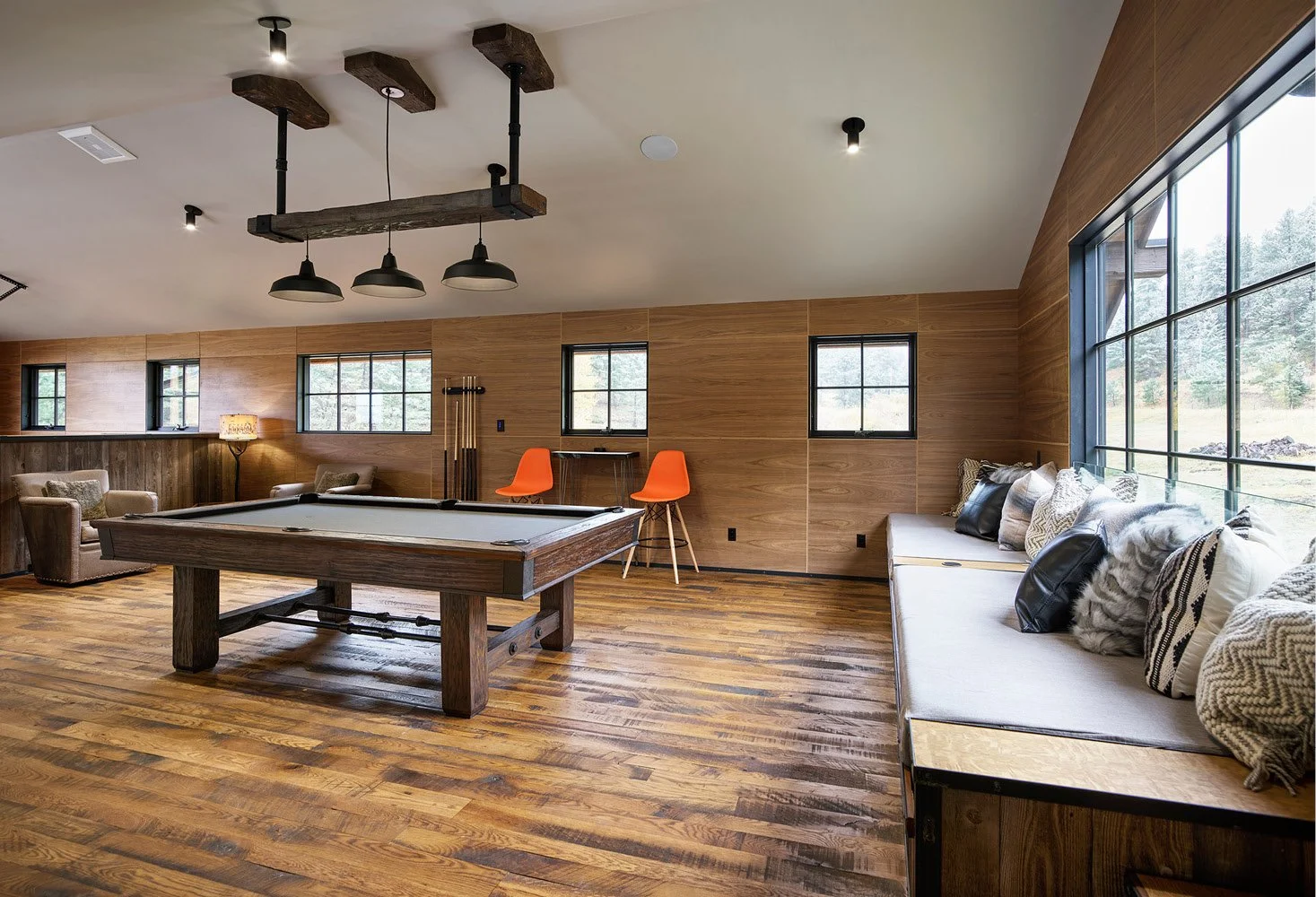 A spacious game room with wood-paneled walls and hardwood floors, featuring a pool table, corner seating with armchairs and a sofa with pillows, a large window seat with pillows, and modern lighting fixtures.