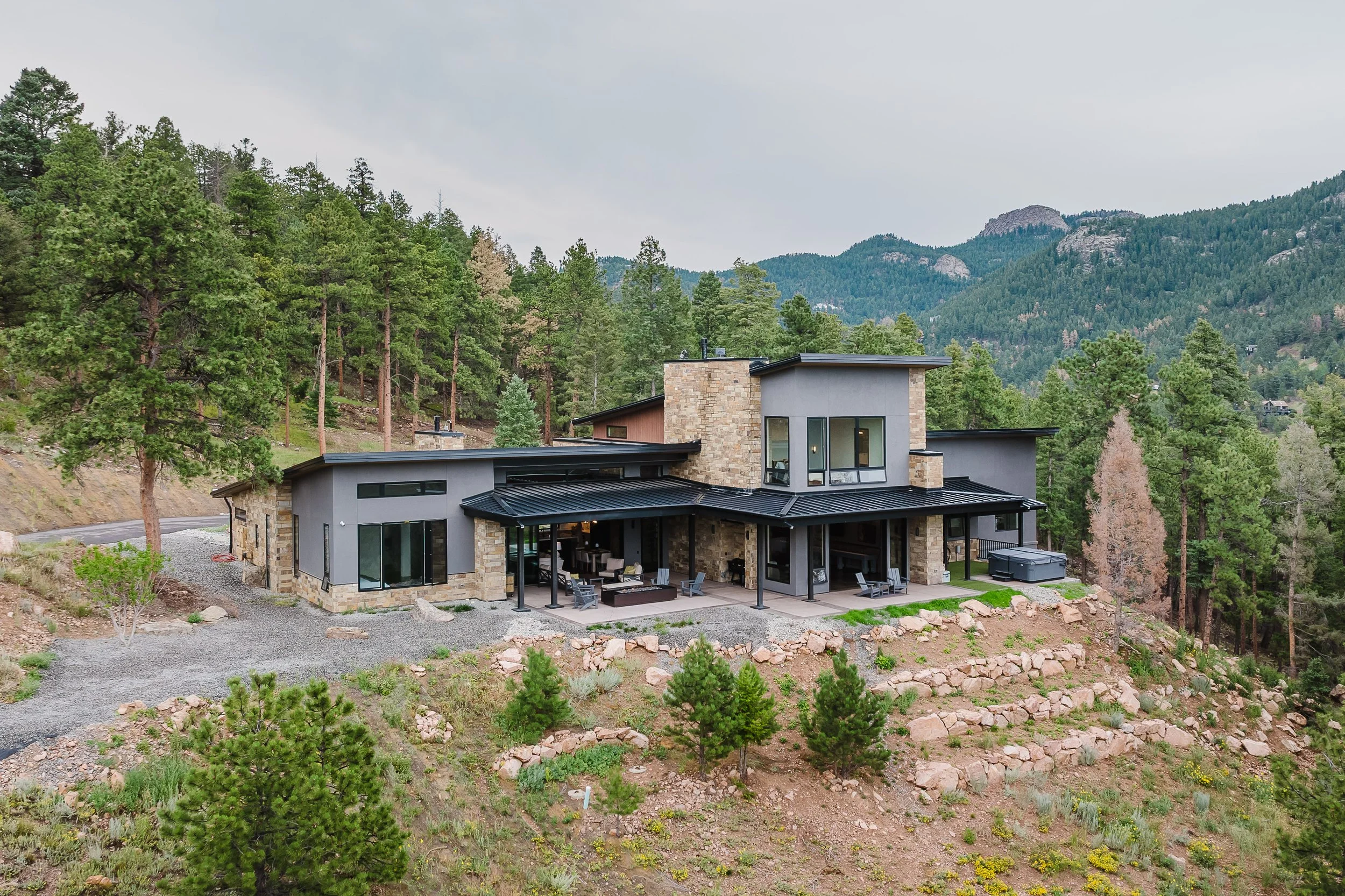 Modern house with stone and gray exterior walls, large glass windows, and a black metal roof, situated on a hillside surrounded by pine trees and mountains in the background.