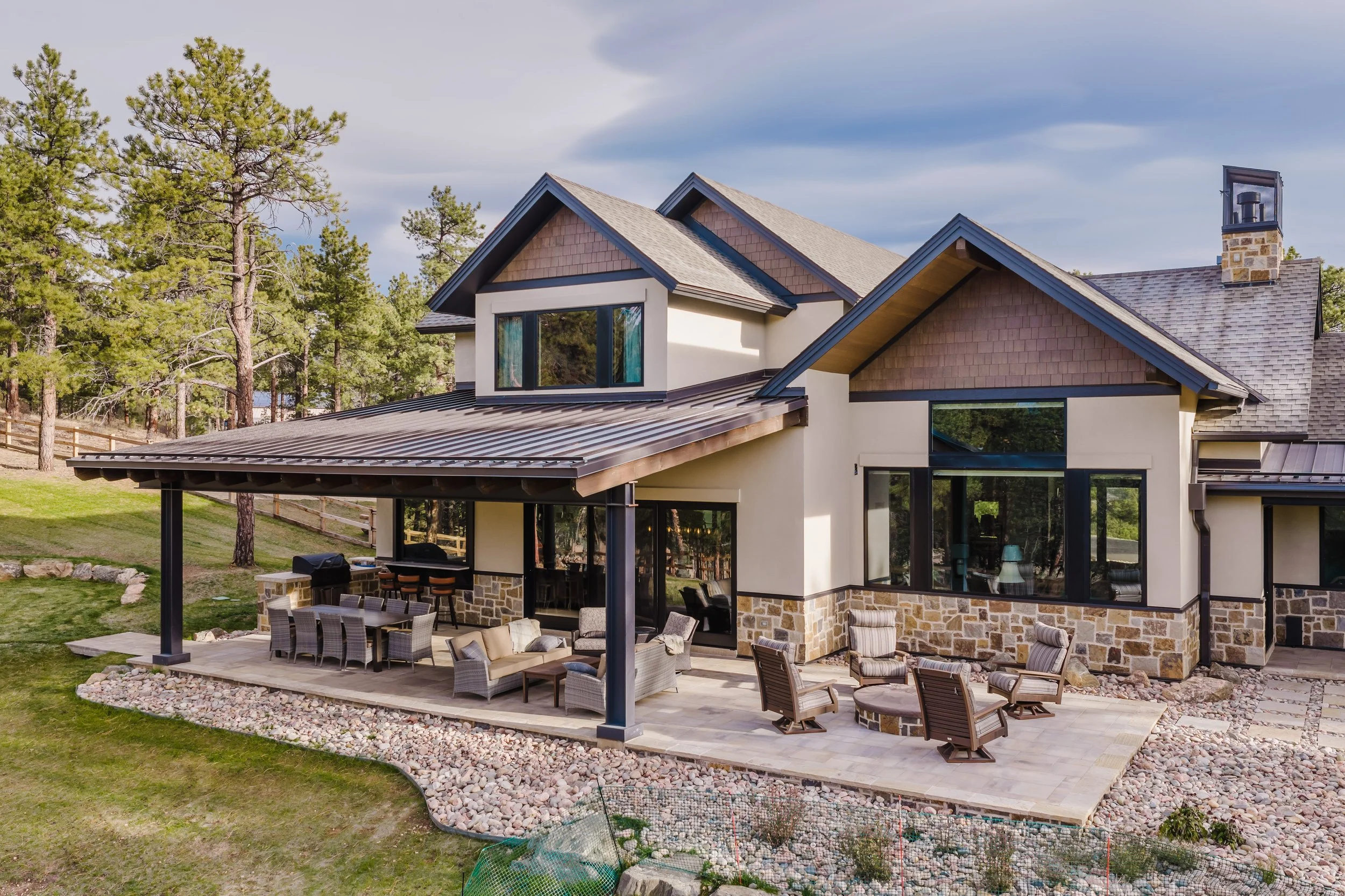 Modern house with a stone exterior and large windows, surrounded by pine trees, with a spacious outdoor patio area with chairs, tables, and a grill.