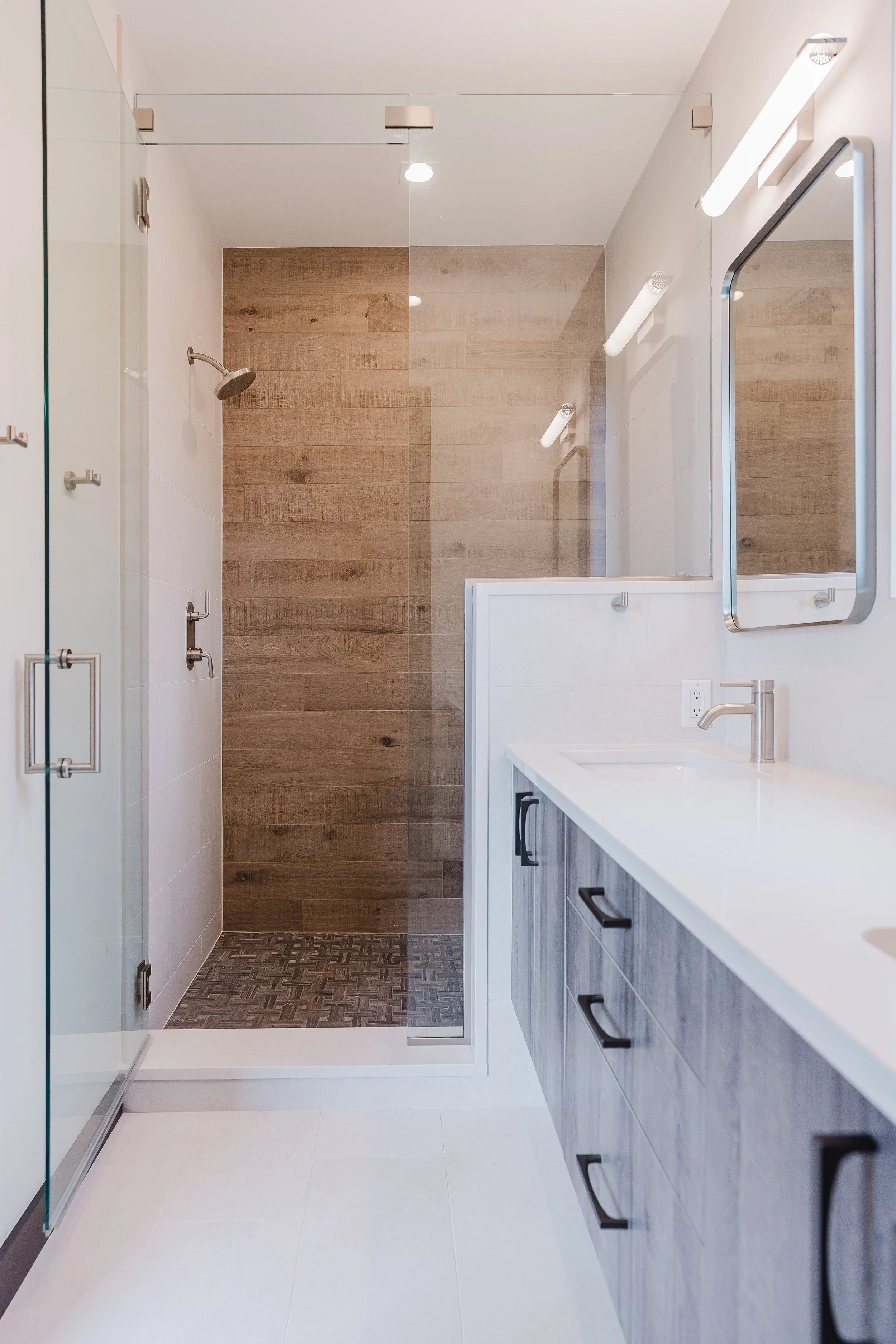 Modern bathroom with walk-in shower area with wood-textured tile walls, a glass partition, a white vanity with black cabinet handles, a rectangular mirror, and wall-mounted lights.