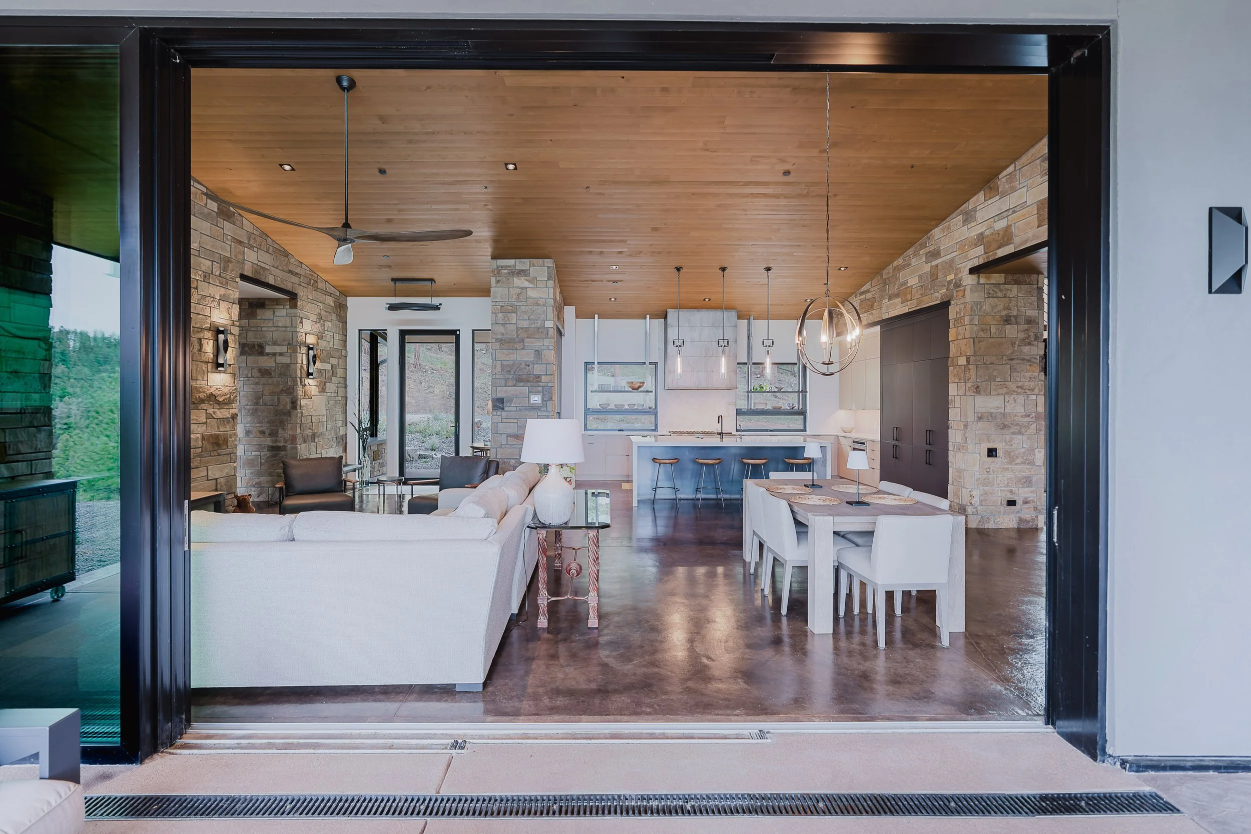 Interior view of a modern open-concept living space with a stone accent wall, wooden ceiling, and large windows, featuring a white sofa, dining table with chairs, and kitchen area with bar stools and pendant lighting.