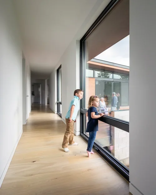 Two children, a boy and a girl, looking out a large window in a modern building hallway.