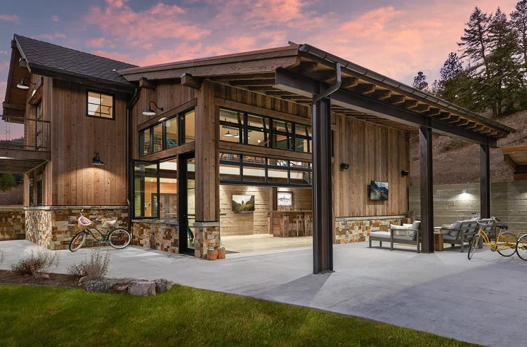 Modern house with a wooden exterior, large glass windows, and an outdoor patio with outdoor seating and bicycles, at sunset.