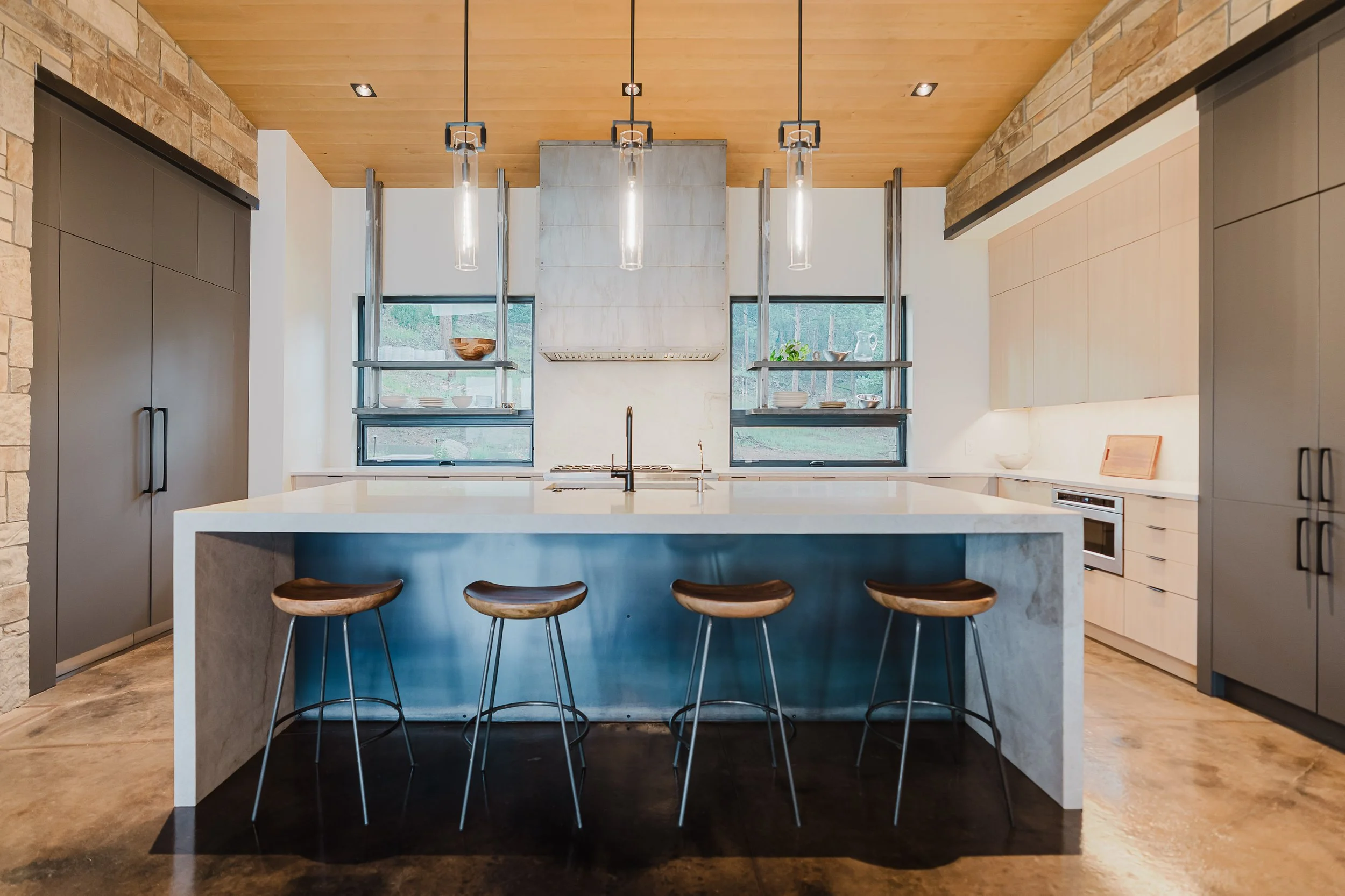 Modern kitchen with a large white island, black bar stools, gray cabinets, and a wood-paneled ceiling, with windows overlooking greenery.