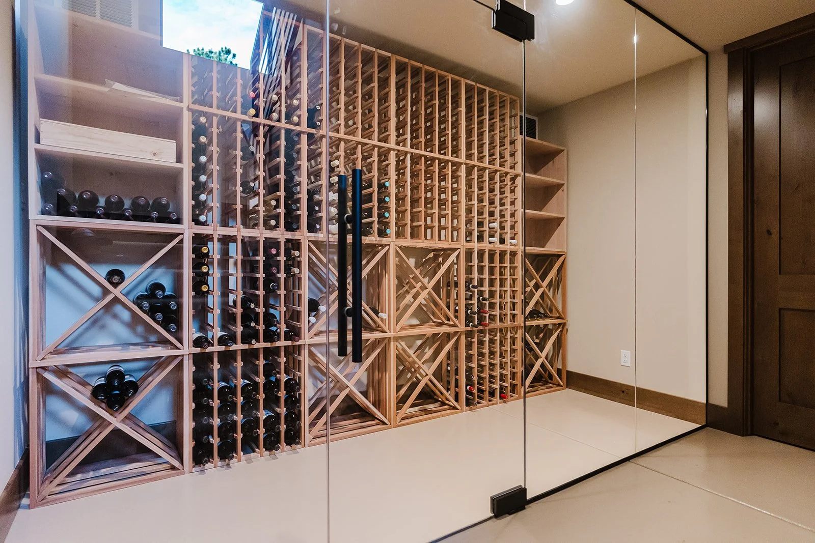 A wine cellar with wooden wine racks holding bottles, behind a glass door with a black handle.