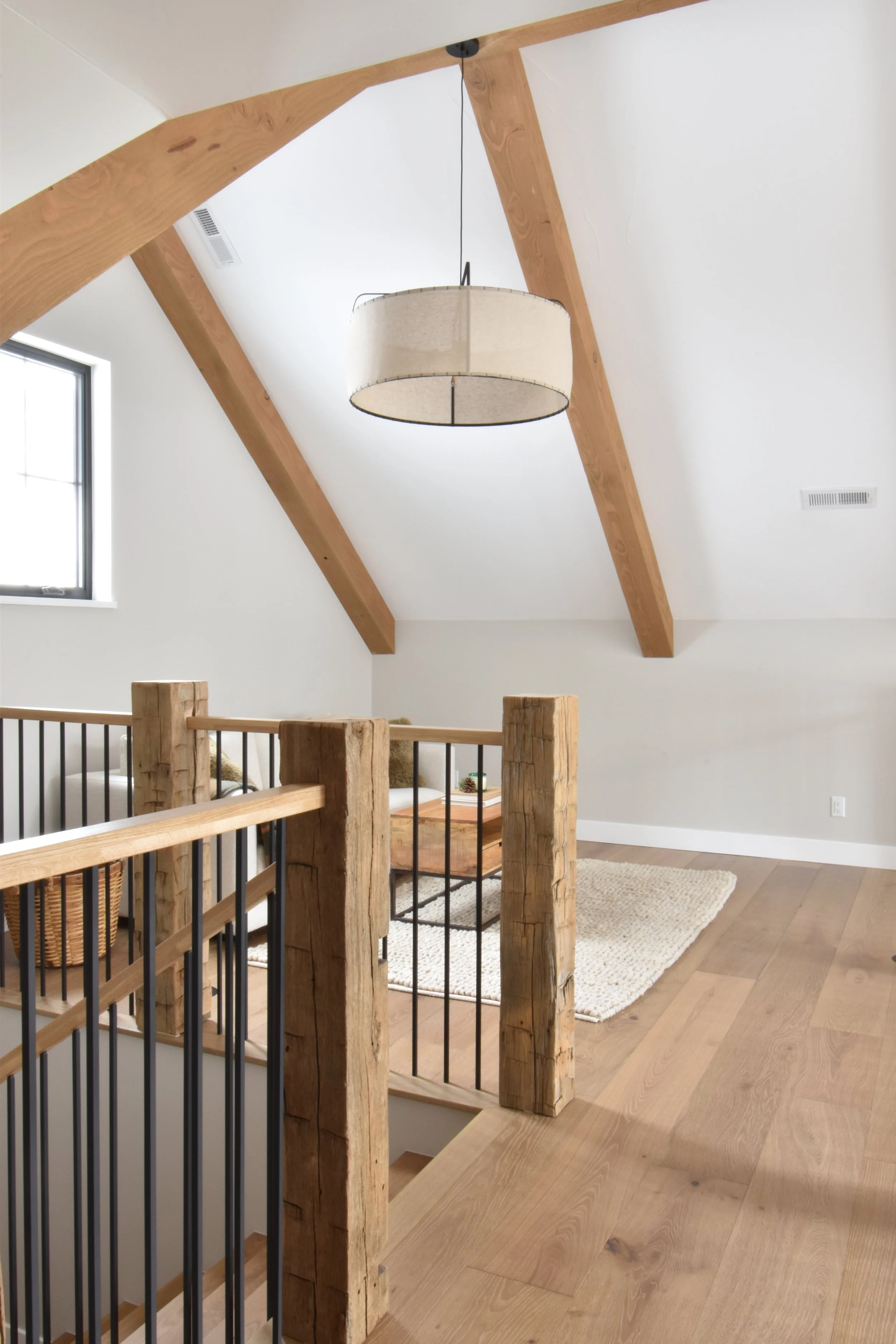 Interior view of a room with a vaulted ceiling featuring wooden beams, a hanging light fixture, a small window, a wooden staircase railing, a white area rug, and hardwood flooring.