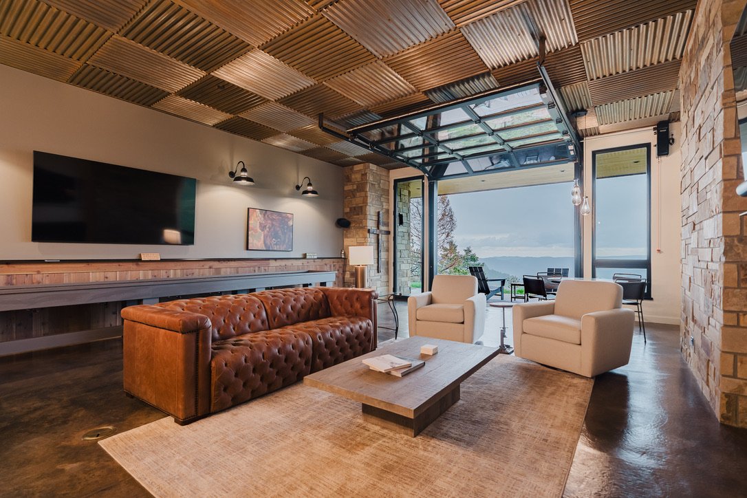 Living room with a leather tufted sofa, two white armchairs, a wooden coffee table, a large flat-screen TV, and a view through large windows to the outdoors with trees and mountains.