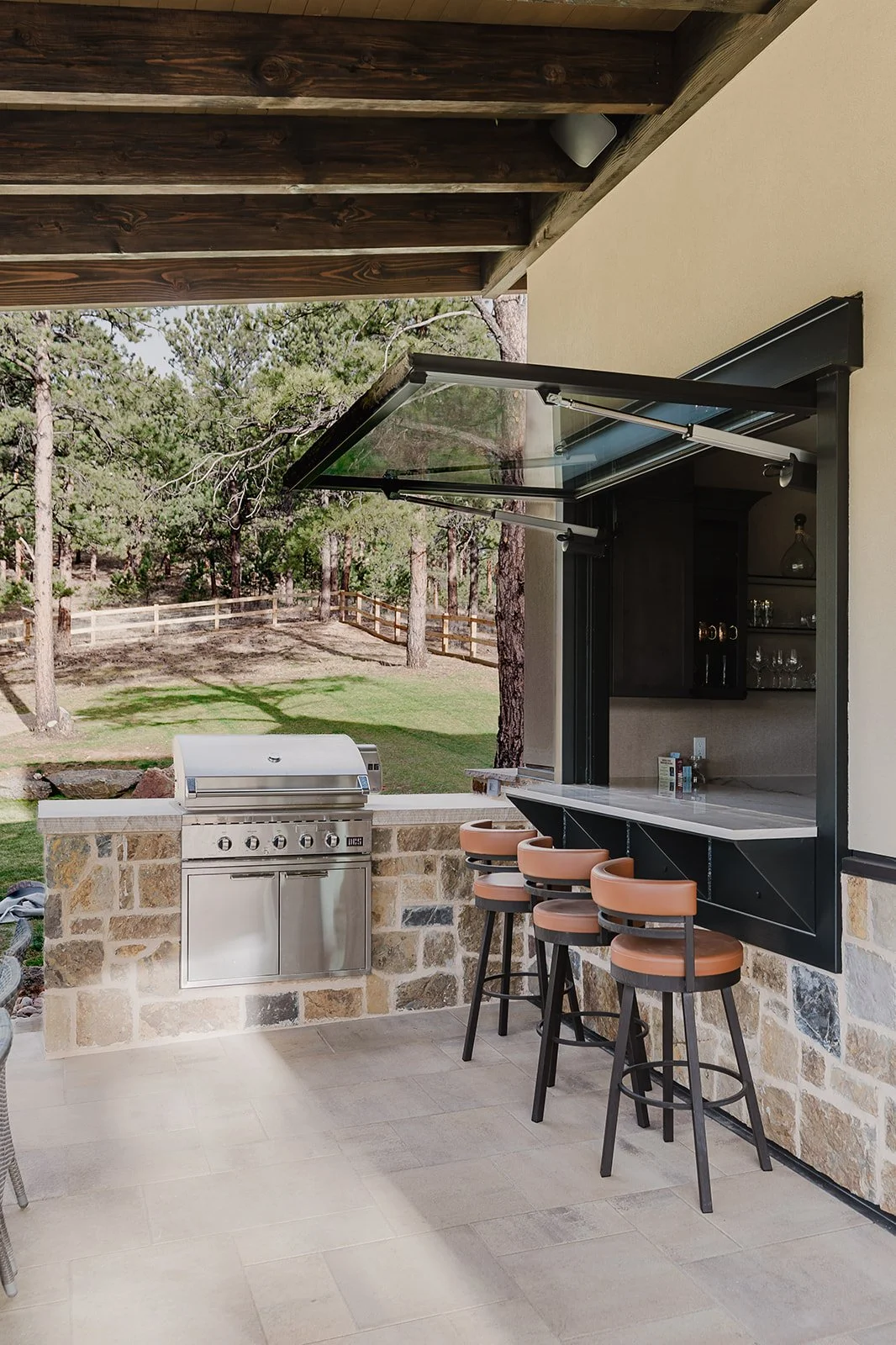 Outdoor patio area with a built-in grill, stone wall, bar with three high chairs, and open window leading to a view of trees and a grassy yard with a wooden fence.