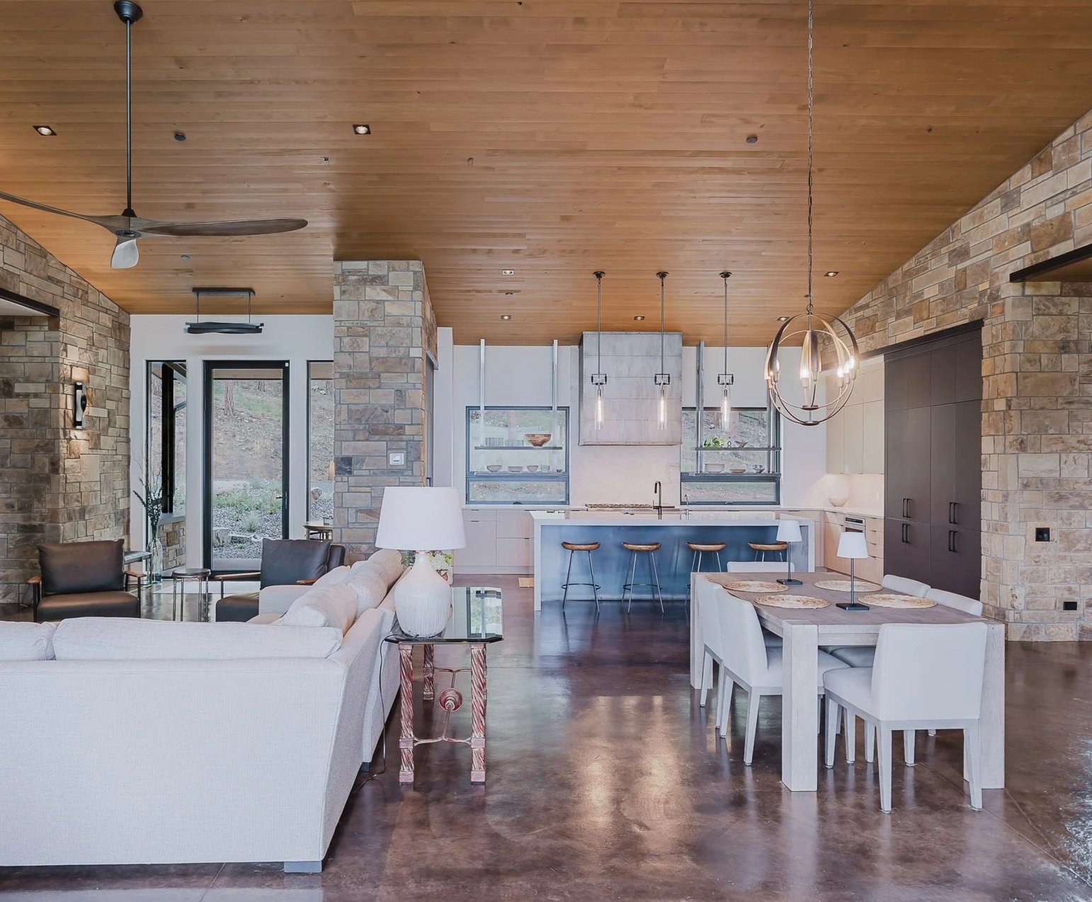 Open-concept living and dining area with wood ceiling, stone walls, and modern furniture including a white sofa, black armchairs, a wooden dining table with white chairs, and a kitchen with a white island and bar stools. Large windows and glass door provide natural light.