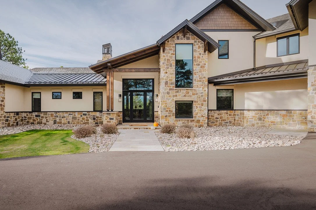 Front view of a modern house with stone and beige siding, black windows, and a metal roof. There is a concrete pathway leading to the black front door with plants and rocks in the landscaping.