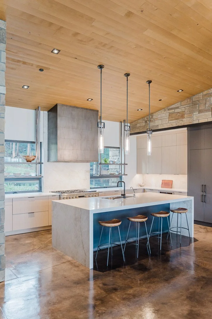 Modern kitchen with white island, three wooden stools, pendant lights, concrete and wooden ceiling, stone and white cabinetry, and large windows.