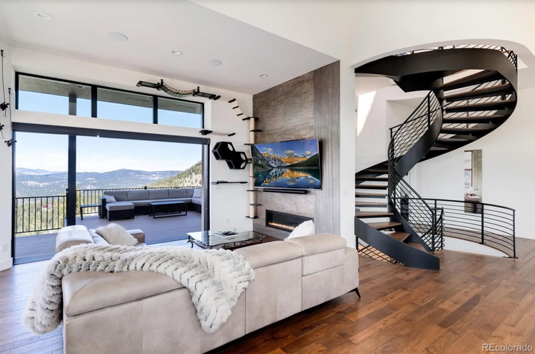 Modern living room with large window overlooking scenic mountain view, beige sectional sofa with furry throw, wall-mounted TV, and spiral staircase with black railing.