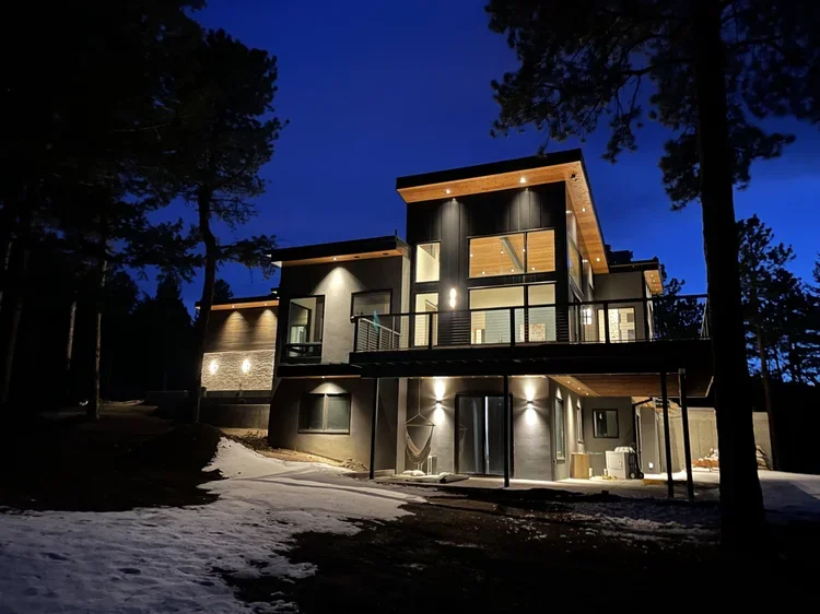 Modern multi-story house with large windows, exterior lighting, a balcony, and a sloped roof, surrounded by trees at night with patches of snow on the ground.