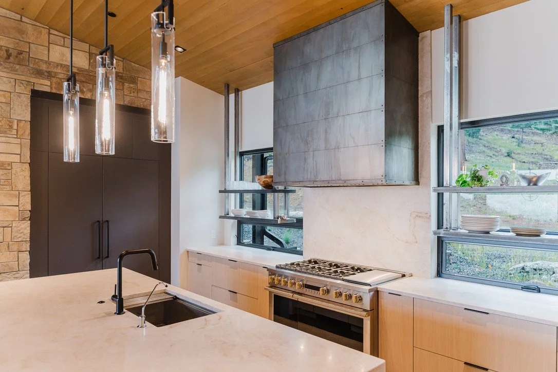 Modern kitchen with beige marble countertop, black faucet, black cabinets, stainless steel stove, and open shelving near windows with dishes and greenery.