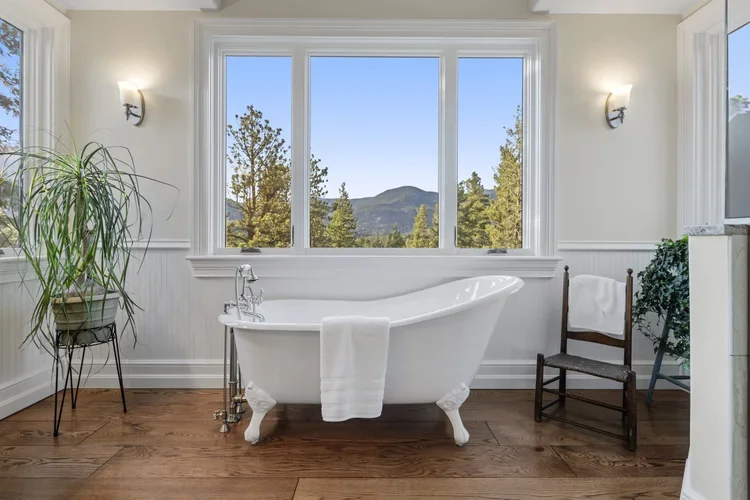 A bright bathroom with a white clawfoot bathtub facing a large window with a scenic mountain view, wooden floor, potted plant on a stand, and a wooden chair.