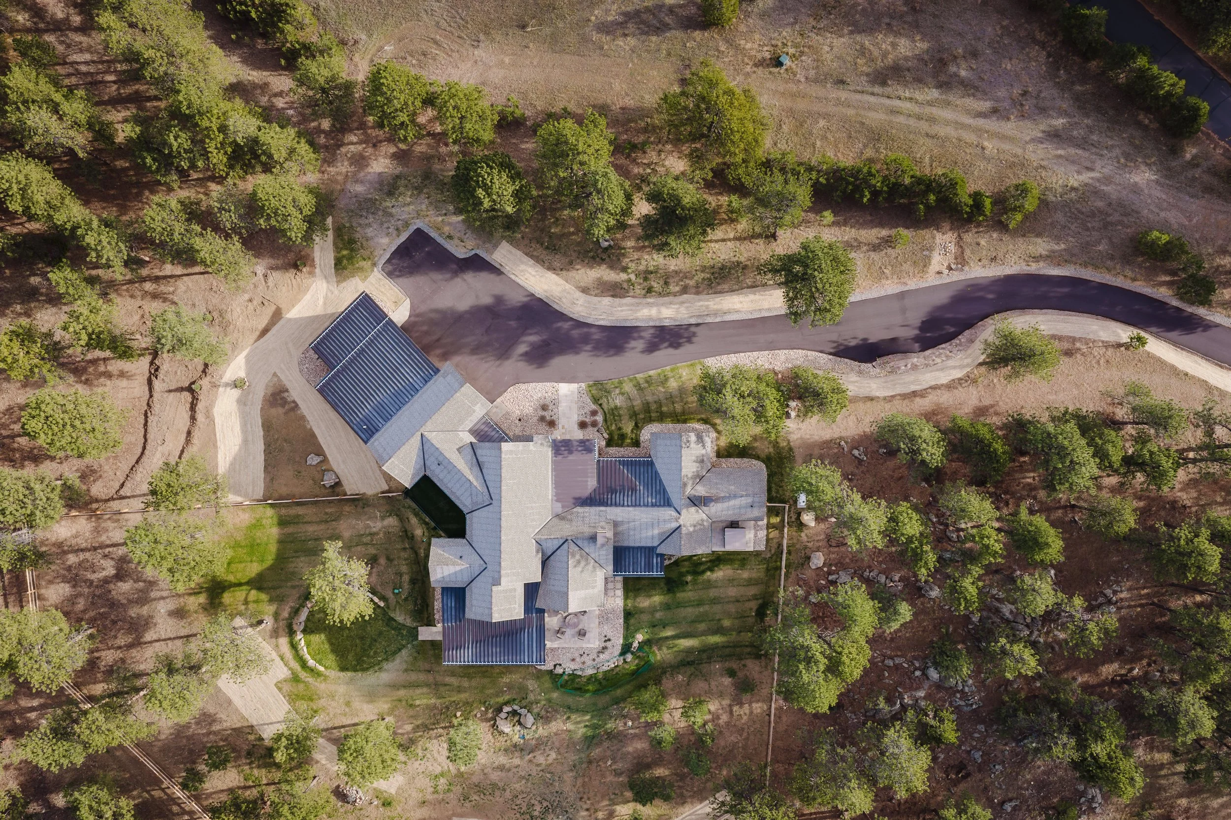 An aerial view of a house with solar panels on its roof, surrounded by numerous trees, a paved road with a curve, and open land with patches of grass and dirt.
