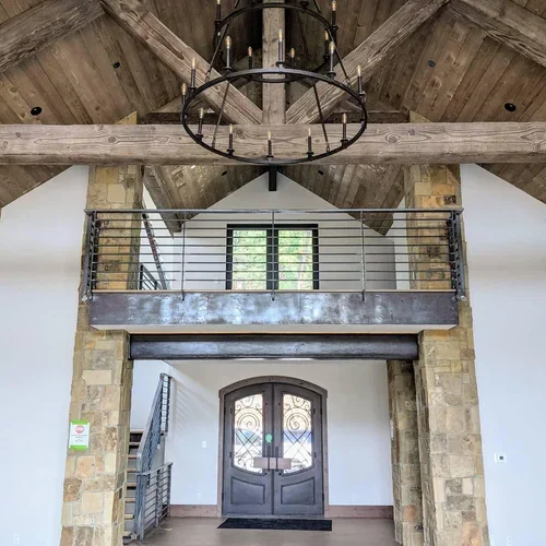 Interior view of a rustic home entryway with a double door, stone columns, and a loft with a metal railing, exposed wooden beams, and a circular chandelier.