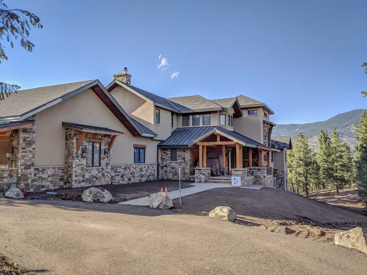 A large new house under construction with stone and stucco exterior, a covered porch supported by wooden beams, and a driveway with dirt and rocks around it, situated in a mountainous area with trees and hills in the background.