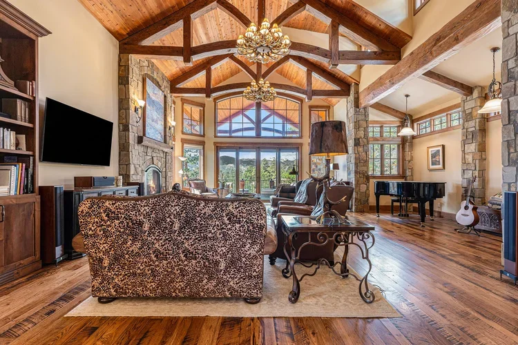 Spacious living room with wooden vaulted ceiling, large windows, stone fireplace, comfortable sofas, a flat-screen TV, a grand piano, and musical instruments, with hardwood floors and warm lighting.