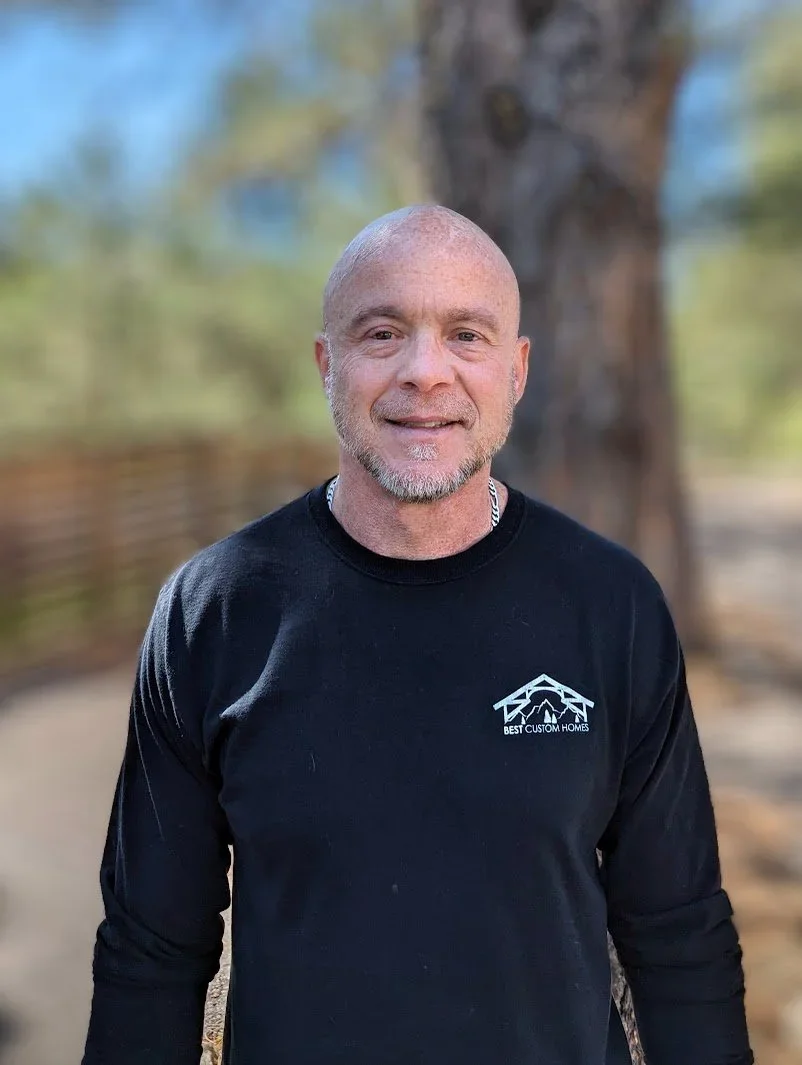 A smiling man with a shaved head and beard, wearing a black long-sleeve shirt with a logo that reads 'Best Custom Homes,' standing outdoors in front of a tree with a blurred background of sky and foliage.
