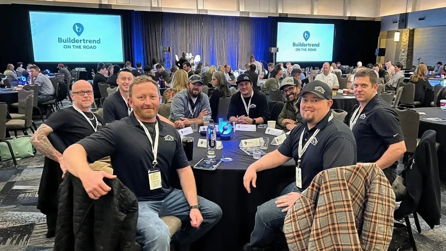 A group of men sitting around a table at a conference, with two large screens in the background displaying the Buildertrend logo and the text "ON THE ROAD." Other attendees are visible in the background.