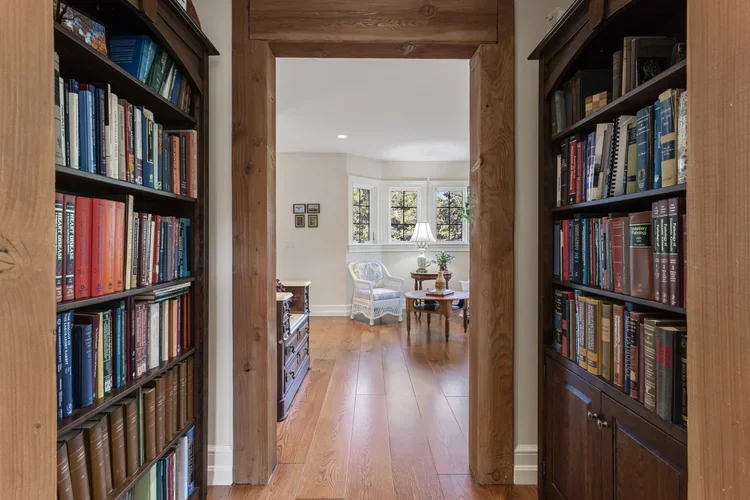 View through a doorway into a bright room with a white armchair, a wooden side table, a lamp, and multiple windows, flanked by bookshelves filled with colorful books.