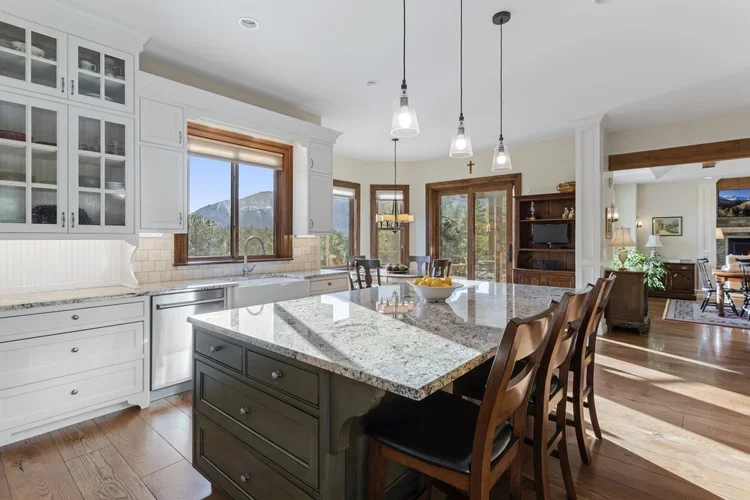 Kitchen with white cabinets, granite countertops, and an island with seating. Large window over the sink showing an outdoor view of mountains. Adjacent dining and living areas with hardwood floors and natural light.