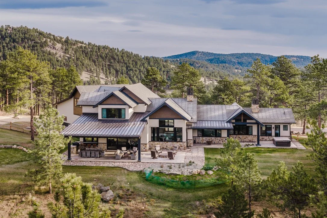 Large modern house with multiple gabled roofs, stone and wood exterior, surrounded by pine trees and mountains in the background, with a landscaped yard and patio area.