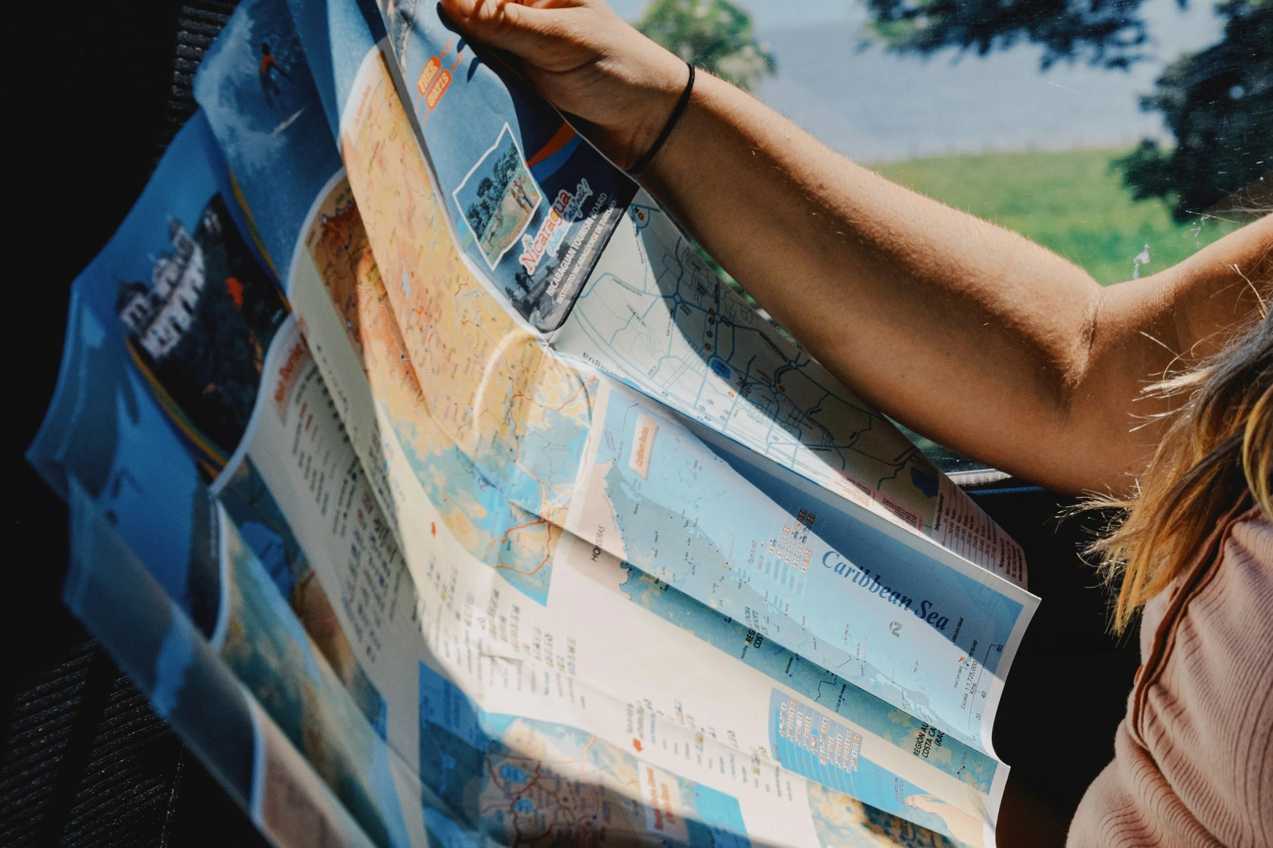 Person holding a travel brochure with a map and information about the Caribbean Sea, with a background of green trees and water.