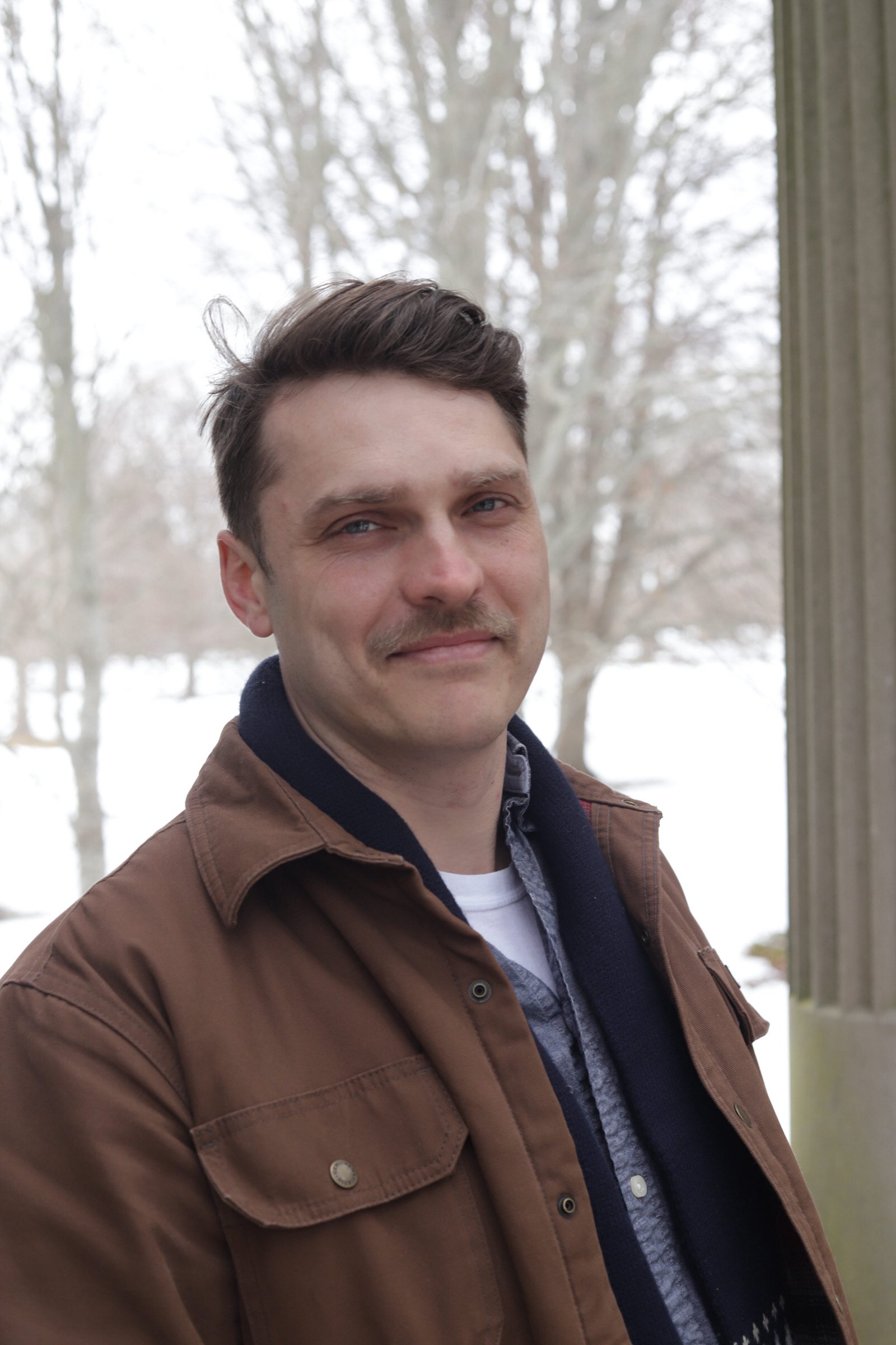 A man with light skin, brown hair, and a mustache standing outdoors on a cloudy winter day with snow on the ground and leafless trees in the background, wearing a brown jacket over a hoodie and a shirt.