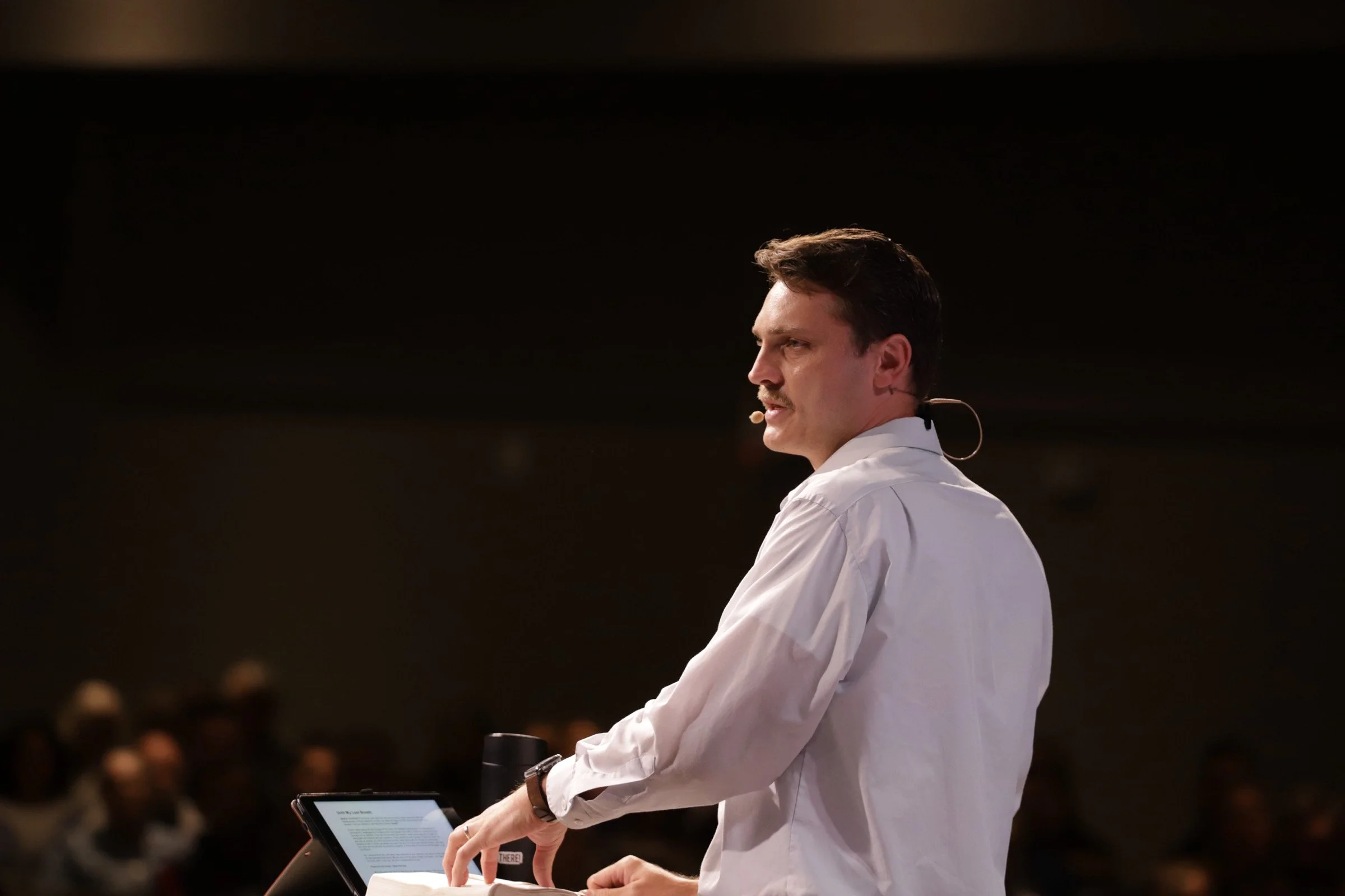 A man in a white shirt giving a presentation on stage, wearing a headset microphone, with an audience in the background.