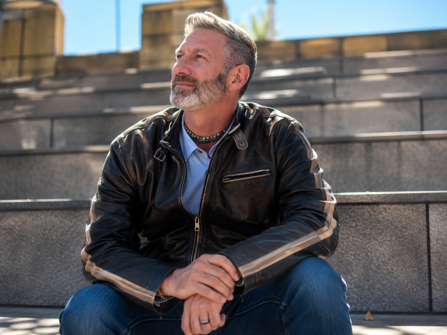 Daniel in leather racing jacket and blue shirt, sitting at a small outdoor ampitheatre on a sunny day...looking at sky