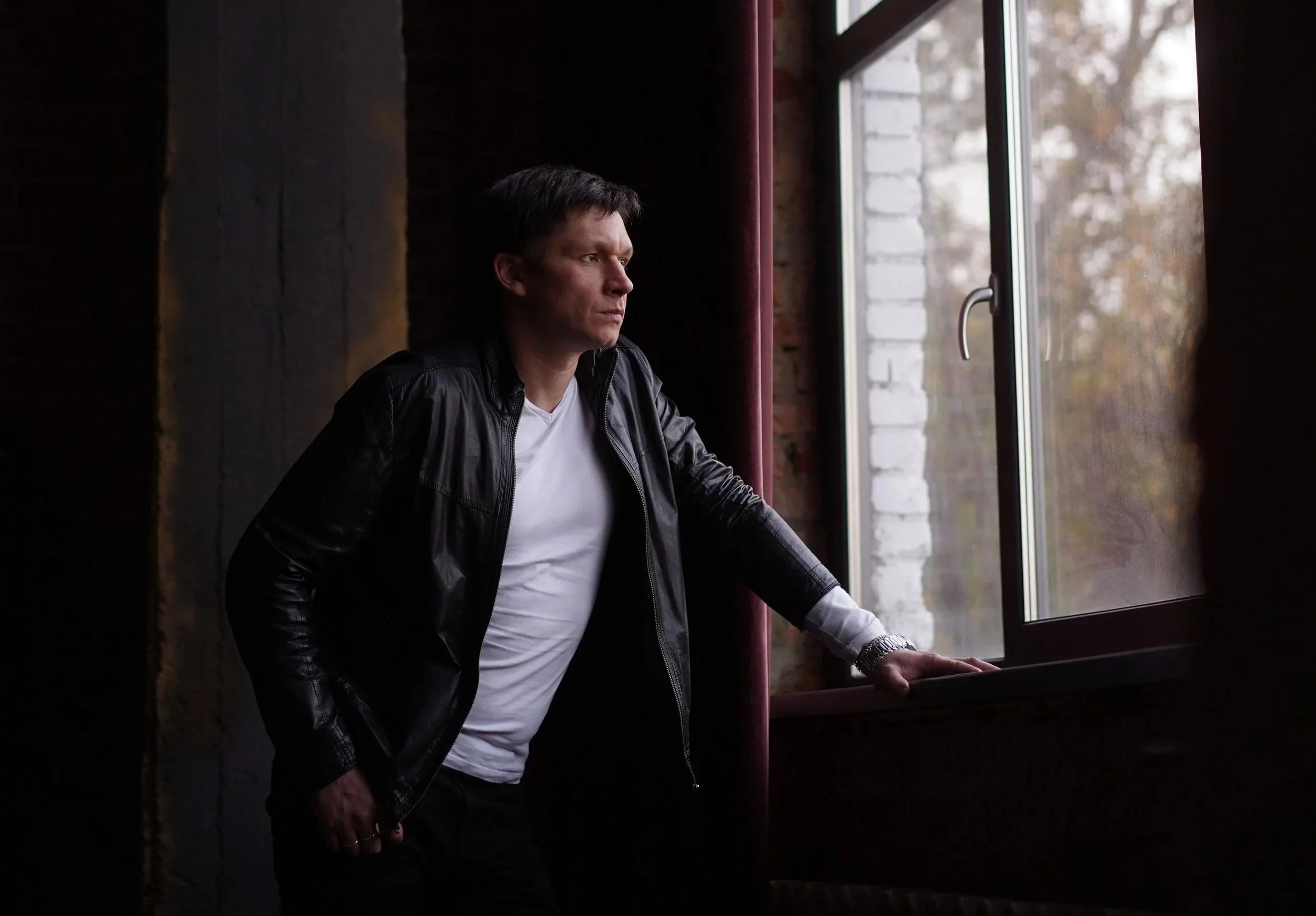 Man in leather jacket and white t-shirt staring out window of darkened room. Looking outside...comtemplation
