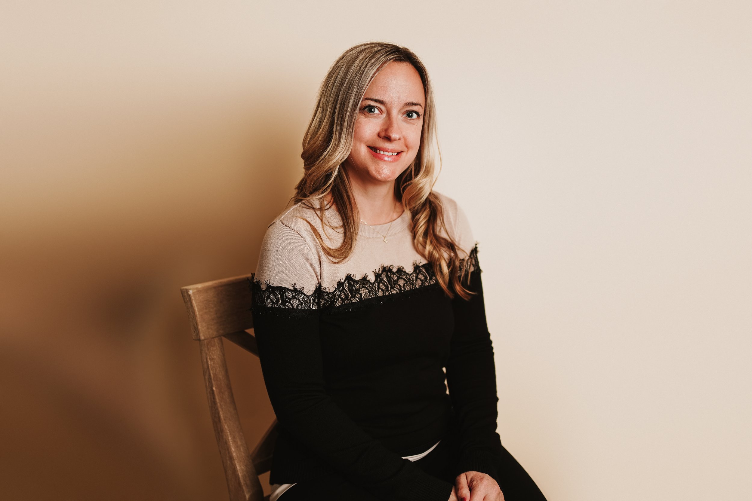 A woman sitting on a wooden chair against a beige wall, wearing a black and beige top with lace details, smiling at the camera.