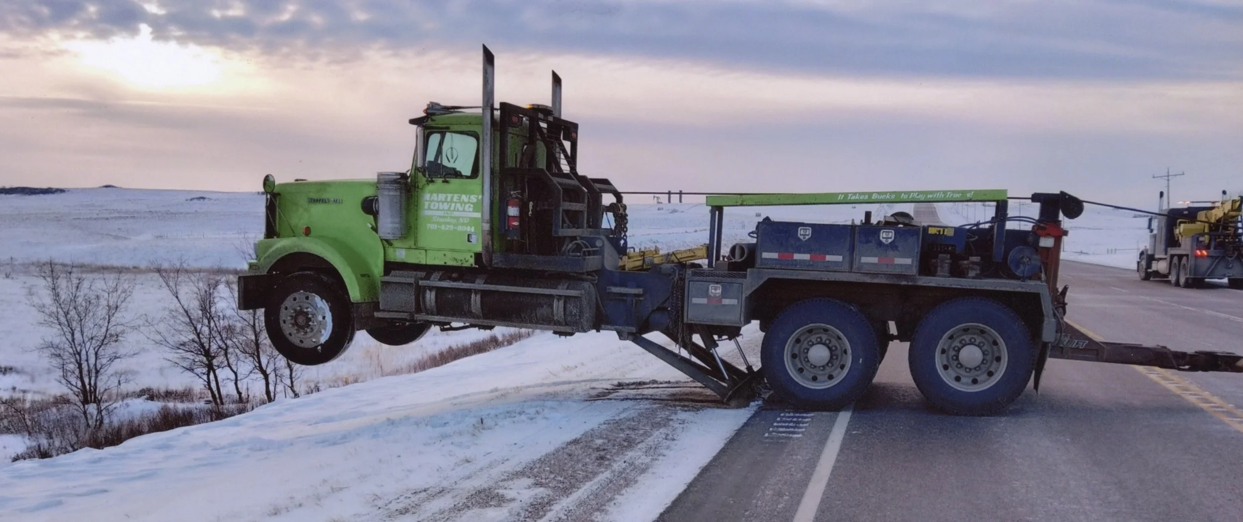 A tow truck with its front wheels lifted off the ground, positioned on a snow-covered roadside with a sunset in the background, and another truck visible further down the road.