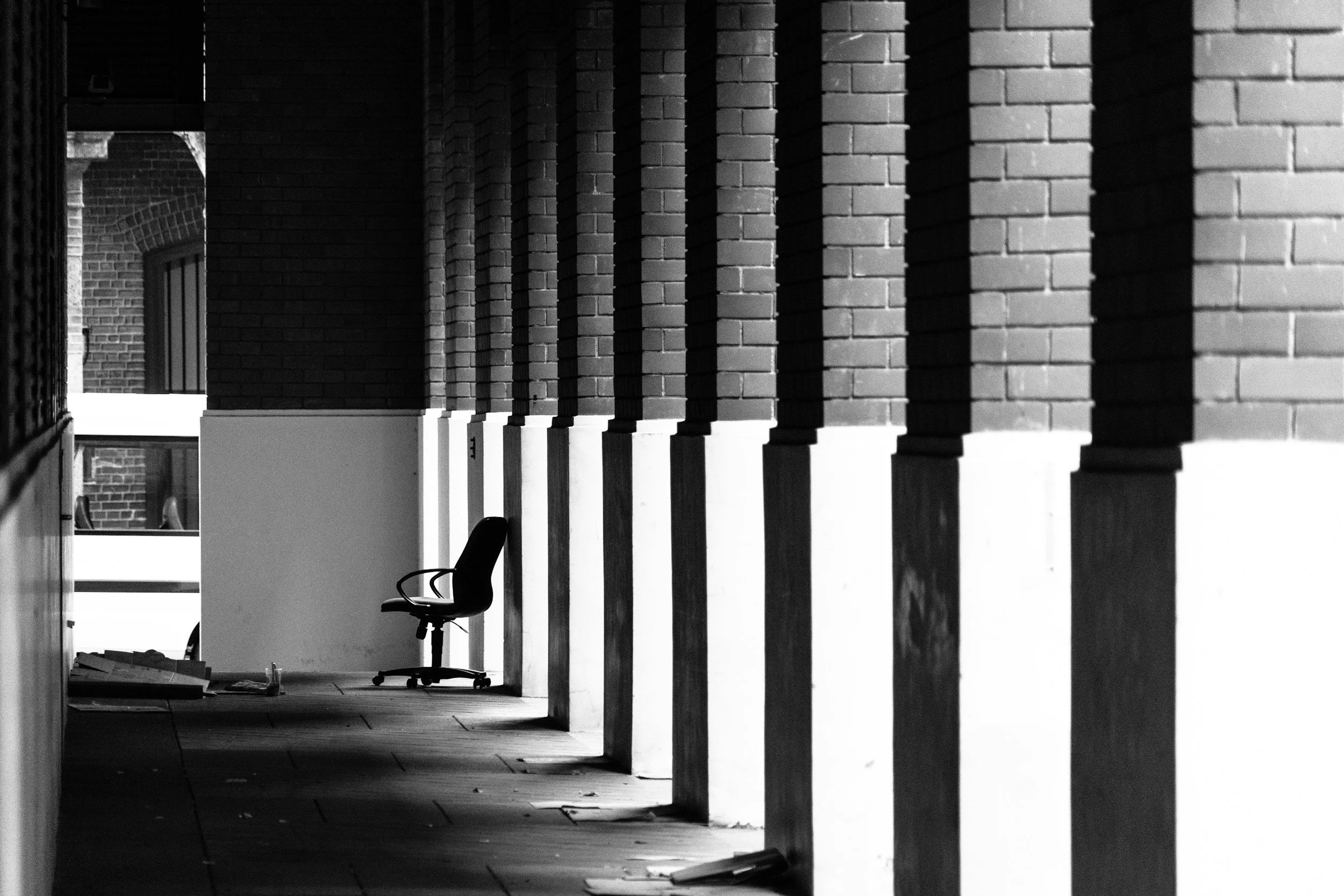 Black and white photo of outdoors corridor with brick columns and an office chair at the end.