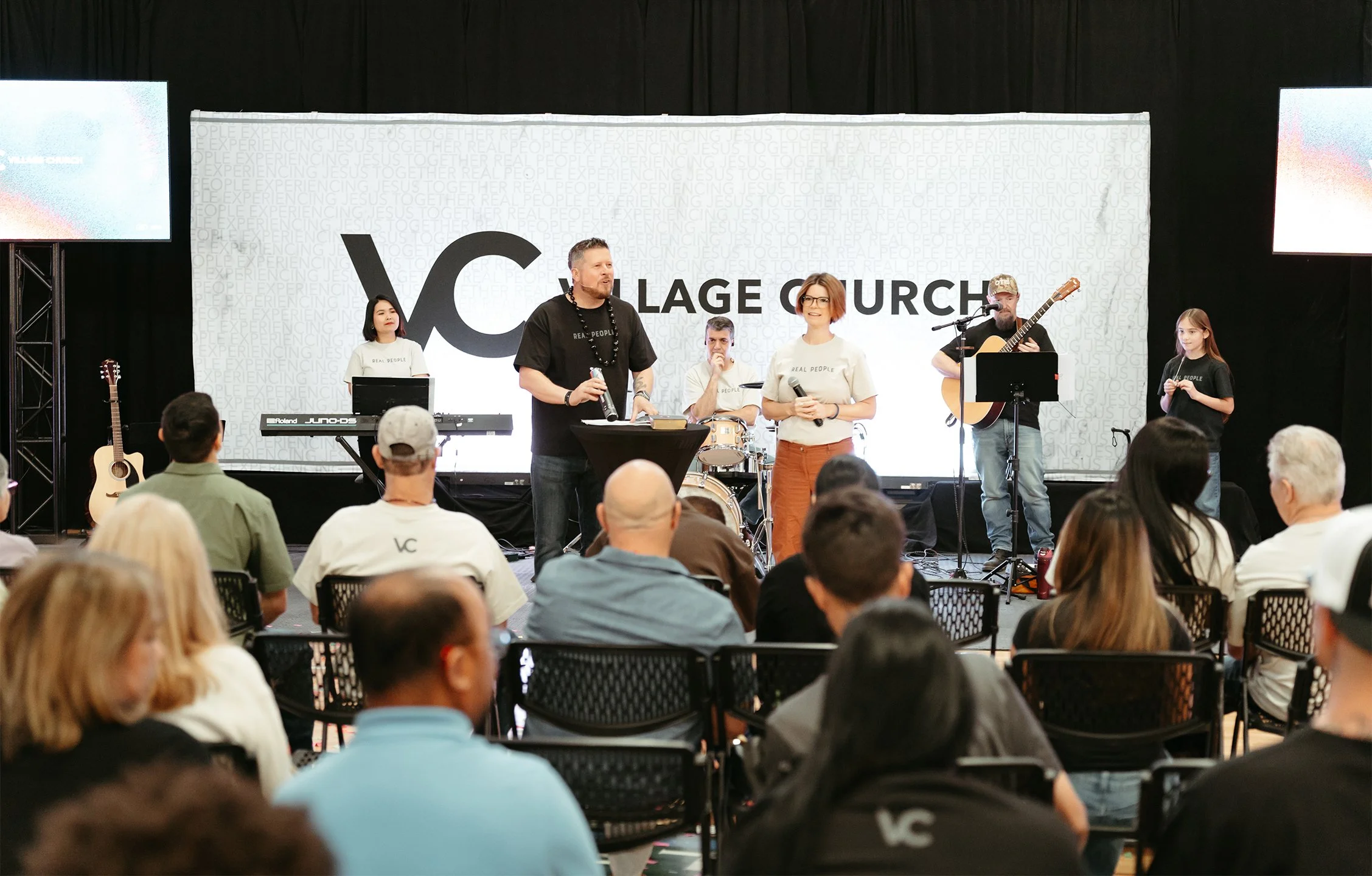 A group of people on stage speaking and performing in front of an audience at a church event, with a large screen displaying a logo behind them.