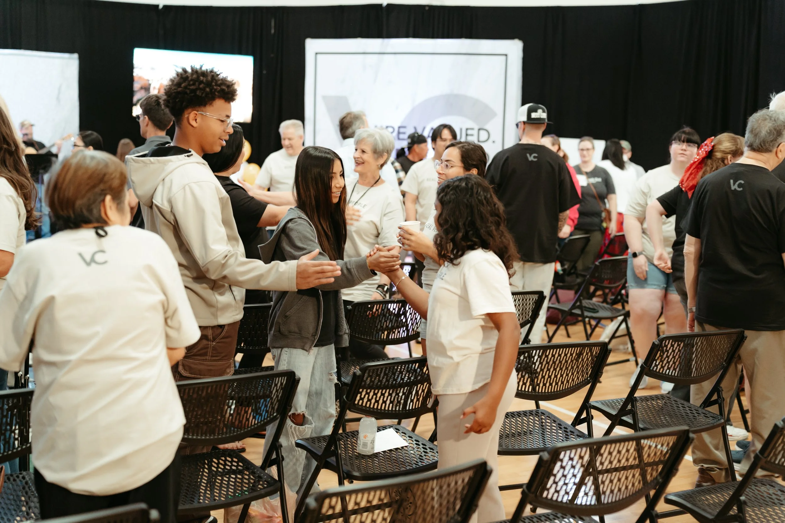 Group of people gathered in a room with black curtains, some sitting and some standing, engaging in conversations, with chairs and tables scattered around.