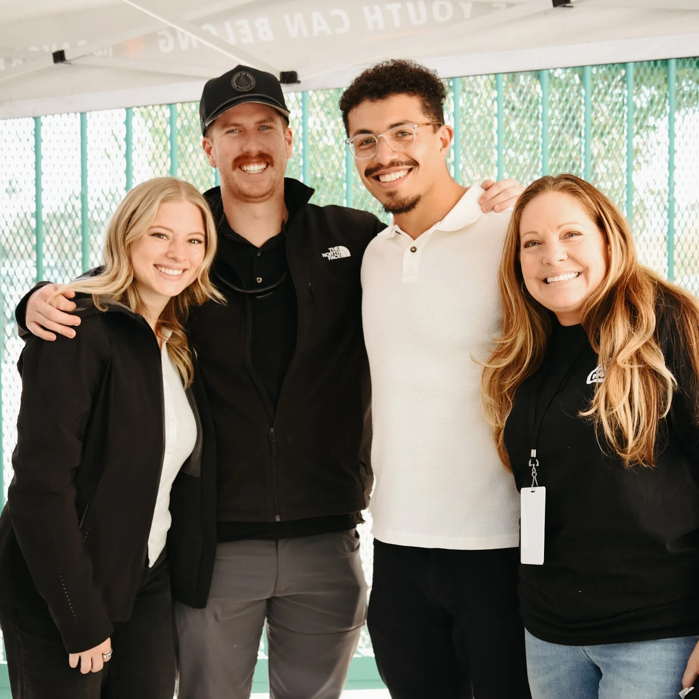 Four people are standing together outdoors, smiling, with two women on the ends and two men in the middle. They are wearing casual clothing.