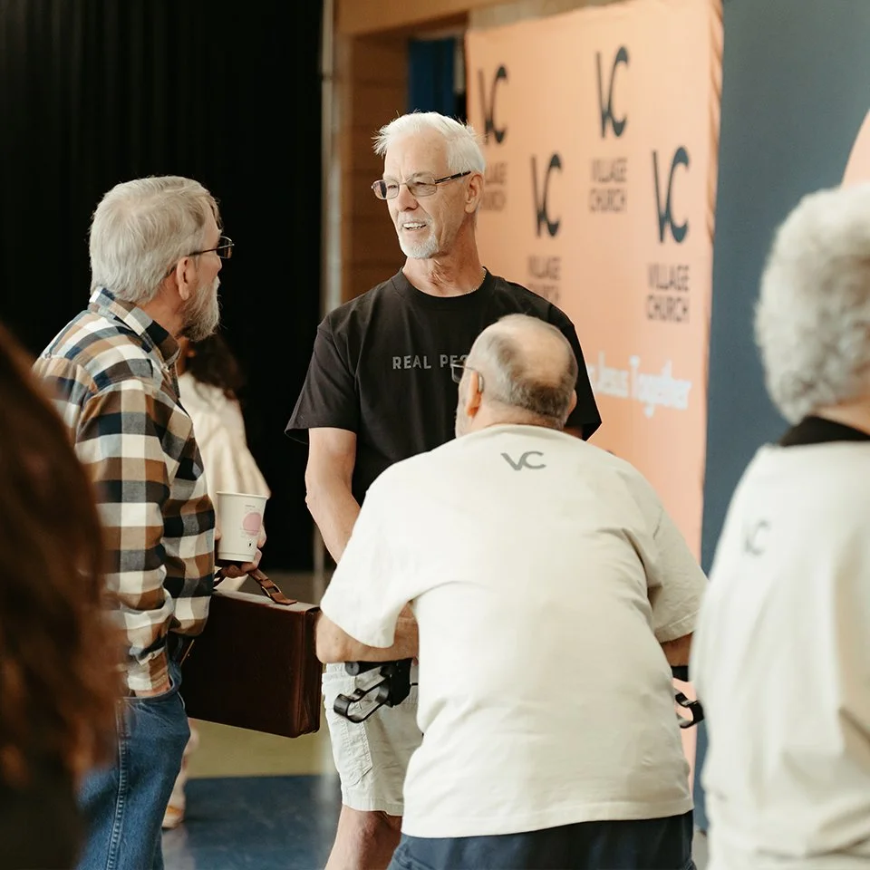 A group of older men engaged in conversation at an event, with a backdrop displaying
